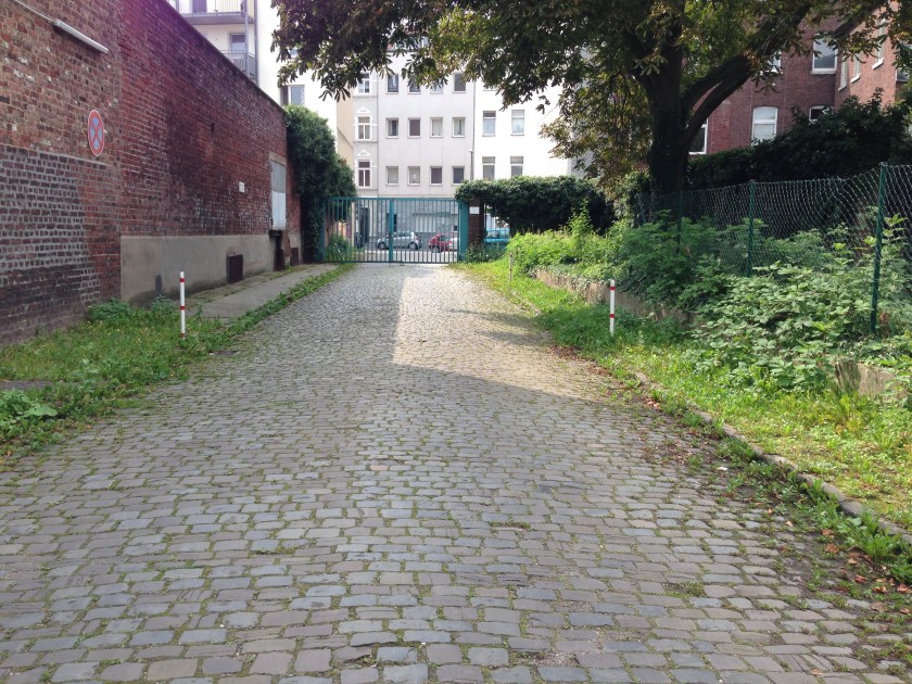 Eine breite Straße führt auf das Gelände der Tuchfabrik Aachen, errichtet 1874 ff. Das alte Pflaster ist erhalten.